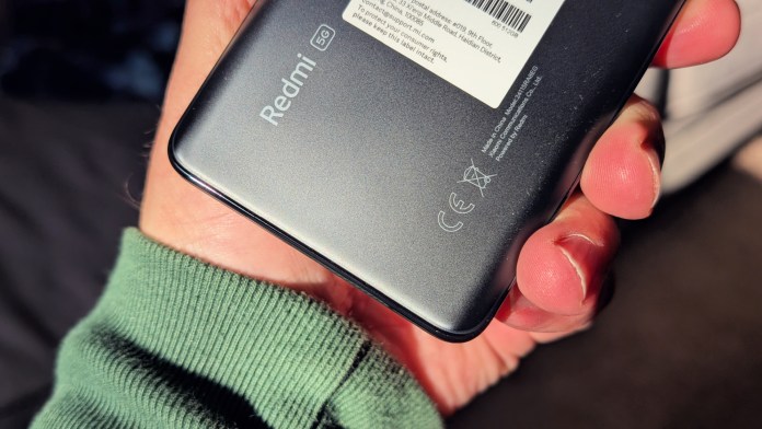 Close-up of the back of a Redmi smartphone, showcasing the 'Redmi' logo, 5G label, and regulatory information, held in a person's hand wearing a green sweater.
