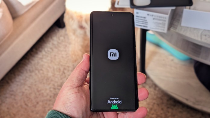 A person holding a Redmi Note 14+ Pro 5G smartphone displaying the MI logo and Android branding on the screen, with a blurred background of a living space.