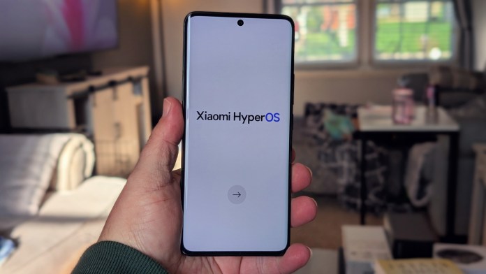 A hand holding a smartphone displaying the Xiaomi HyperOS startup screen in a home setting.