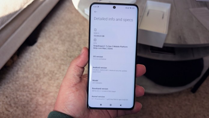 A person holding a smartphone displaying detailed specifications, including RAM, CPU, OS version, and Android version, with a modern interior background.