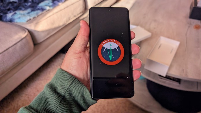 A person holding a smartphone displaying the Android 14 logo on its screen, with a cozy living room setting in the background.