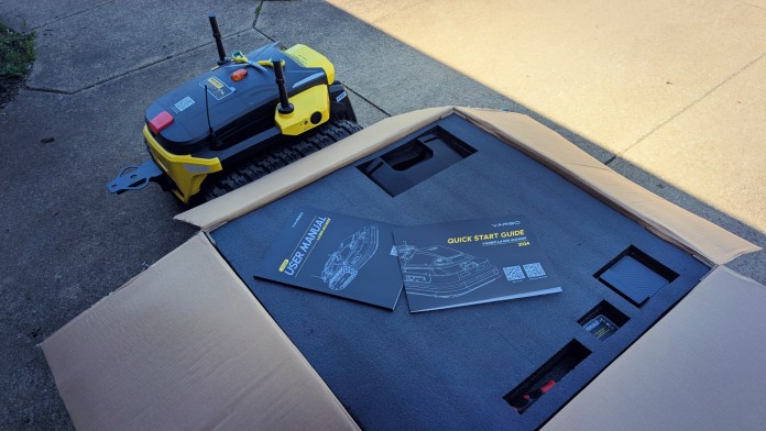 A modular yard robot, the Yarbo Core, alongside its packaging, including a user manual, displayed on a concrete surface.