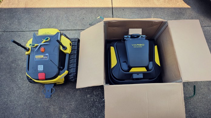 An overhead view of a Yarbo modular yard robot positioned beside an open box containing another module, with both items appearing on a concrete surface.
