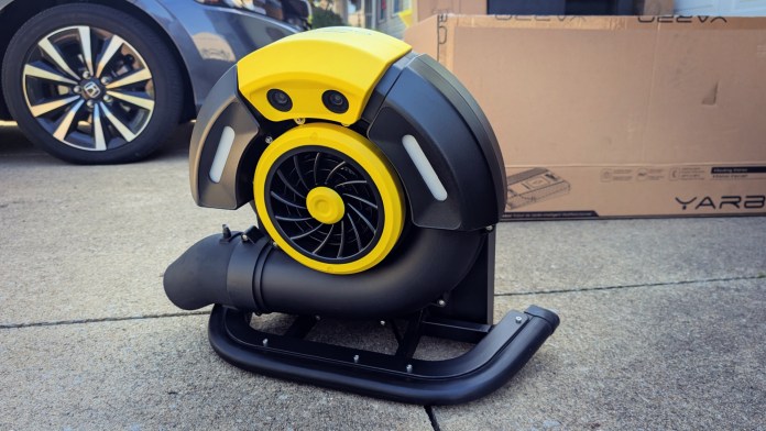 A close-up view of a yellow and black leaf blower module for the Yarbo Core modular yard robot, positioned on a concrete surface with a car and a partially visible box in the background.