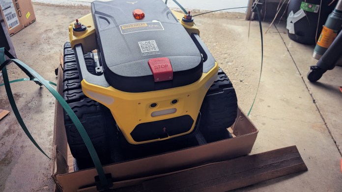 A modular yard robot named Yarbo Core, partially unpacked from its cardboard box, showing its large wheels and control interface in a garage environment.
