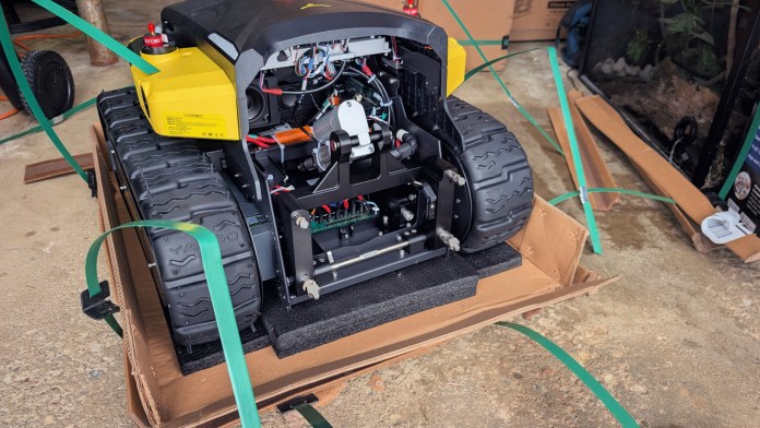 An image of the Yarbo Core modular yard robot showing its internal components and wiring, with straps securing it on a cardboard base in a garage setting.