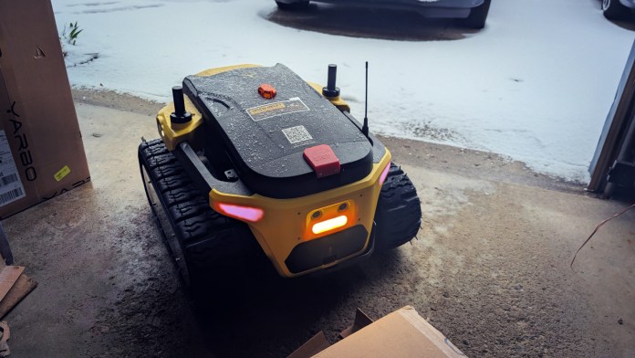 A modular yard robot, the Yarbo Core, sits in a garage with snow visible outside, showcasing its rugged design and readiness for outdoor tasks.