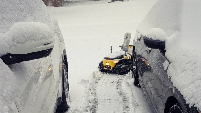 A modular yard robot, the Yarbo Core, clearing snow from a driveway between two parked cars under snowy weather.