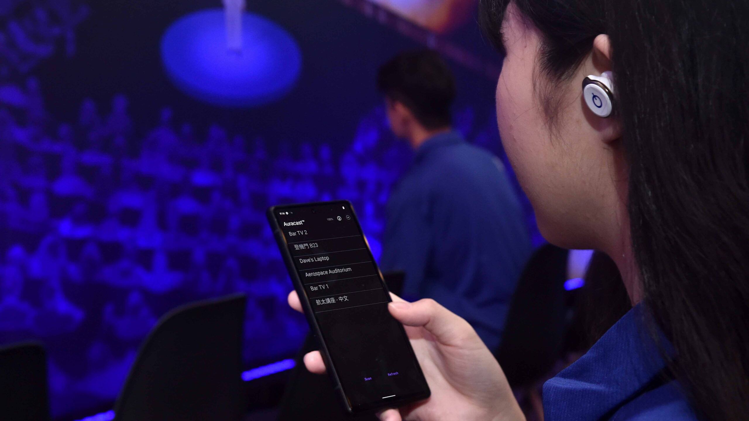 Photo of a person using earbuds with Auracast settings open on their phone against a blue background.