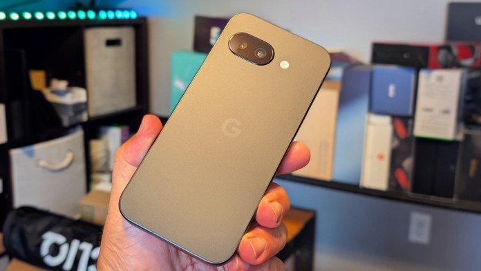 A person holding a Google Pixel 9A smartphone, showcasing its matte-finished back and design, with blurred product packaging in the background.