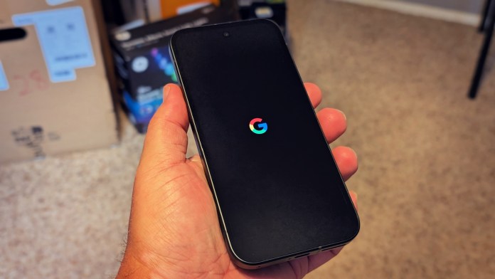 A person holding a Google Pixel 9A smartphone displaying the Google logo on the screen, with boxes and a cluttered background.
