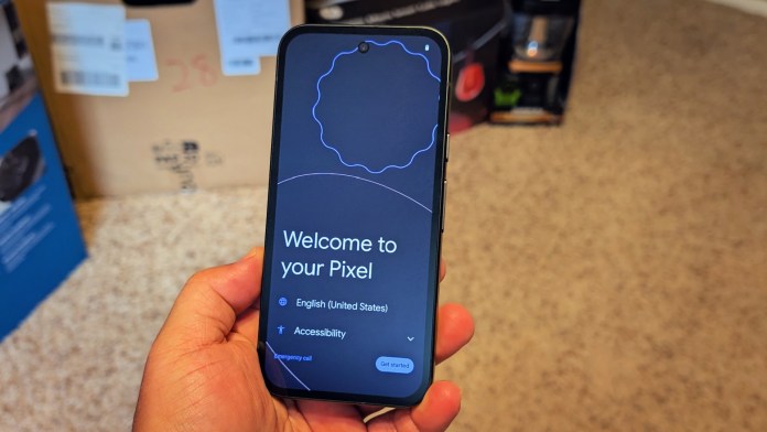 A hand holding a Google Pixel 9A smartphone displaying the welcome screen with options for language and accessibility in a home setting.