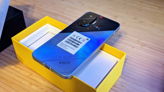 Close-up of the POCO F7 smartphone resting on its packaging. The phone features a unique design with a striking blue and black color scheme, showcasing the rear camera module and branding.