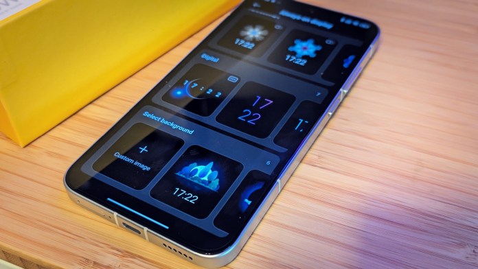Close-up view of the POCO F7 smartphone displaying the customization options for its ambient display, with various digital clock styles and background selections. The phone is positioned on a light wooden surface next to a yellow box.