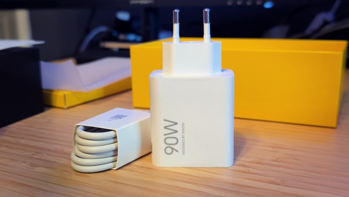A 90W fast charger with two prongs, alongside a coiled USB cable and a yellow packaging box, placed on a wooden surface.