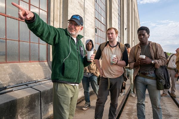 Director Francis Lawrence engages with actors Cooper Hoffman and David Jonsson on the set of 'The Long Walk', with other cast members in the background. All are holding cups, standing in front of a building with large windows.