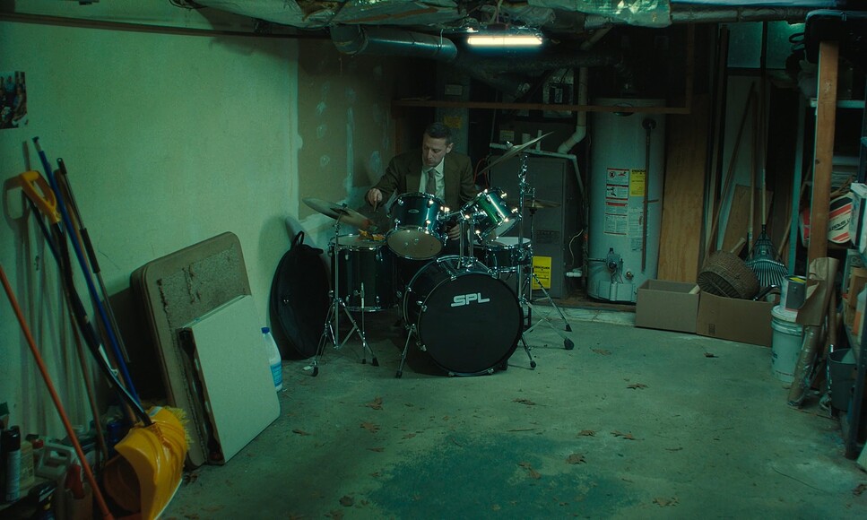 Tim Robinson in 'Friendship' playing a drum kit in an empty garage.