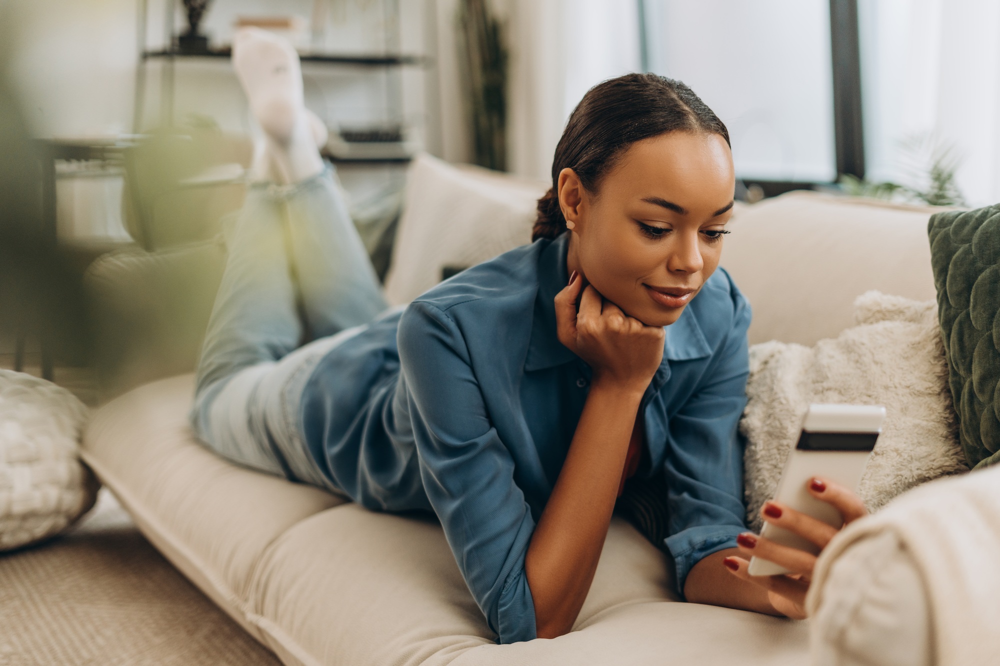 Smiling, African American woman holding mobile phone, social media lying on comfortable sofa