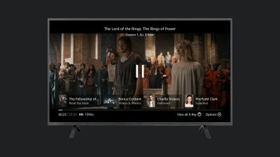 A paused scene from The Lord of the Rings: The Rings of Power on Prime Video shows playback controls and the X-Ray feature. The screen highlights character information, bonus content, and book links at the bottom. A subtle hint suggests pressing the Up button to access the full X-Ray view.