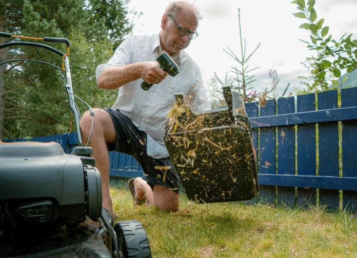 A person in a white shirt and shorts kneels on grass, using a cordless blower to clean up debris from a lawn, with a blue fence and trees in the background.