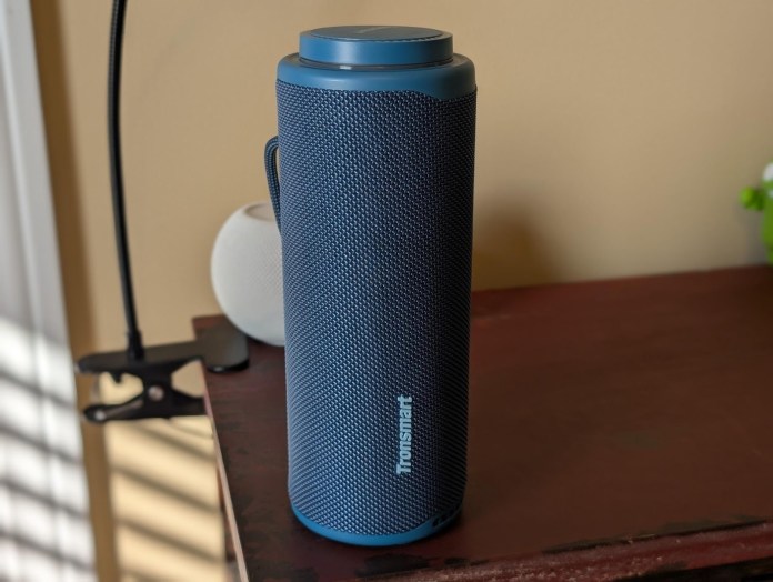 A blue Tronsmart T8 portable outdoor speaker with a textured finish, placed on a wooden surface next to a small white object and a potted plant.