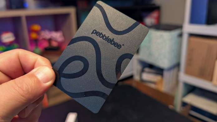 A person holding the Pebblebee Card 5, a slim rechargeable tracker designed to fit into a wallet. The card features a sleek design with the Pebblebee logo and is set against a blurred background of colorful items and storage containers.