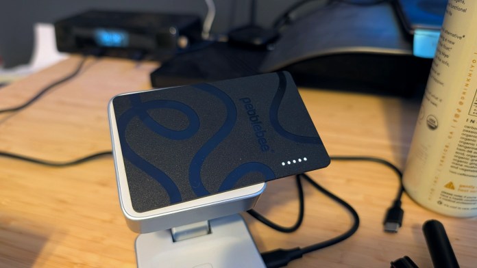 A Pebblebee Card 5 Bluetooth tracker placed on a charging dock, designed to fit in a wallet or card holder, with visible battery indicators.